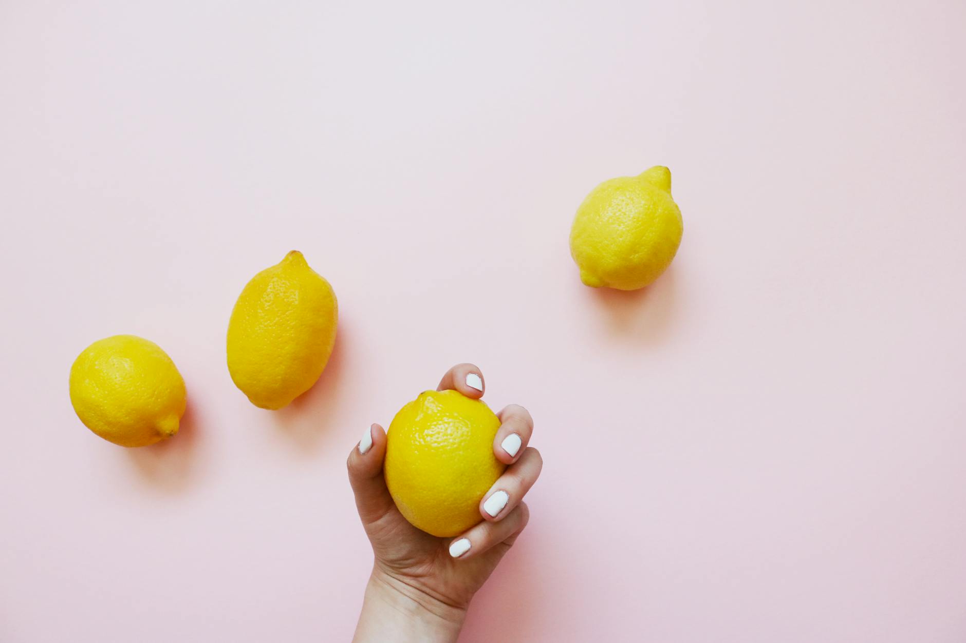 Una mano con uñas blancas sosteniendo un limón fresco sobre un fondo rosa suave, rodeada de tres limones adicionales