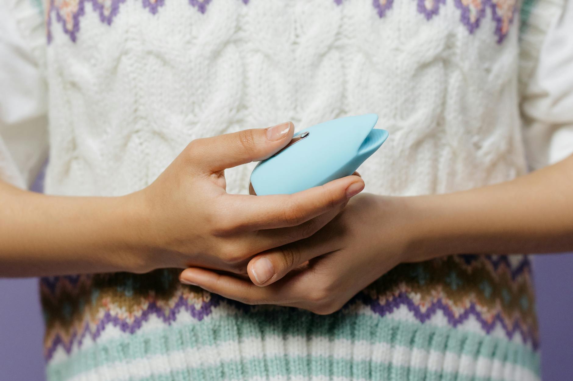Primer plano de manos sosteniendo un vibrador personal azul contra un suéter tejido.